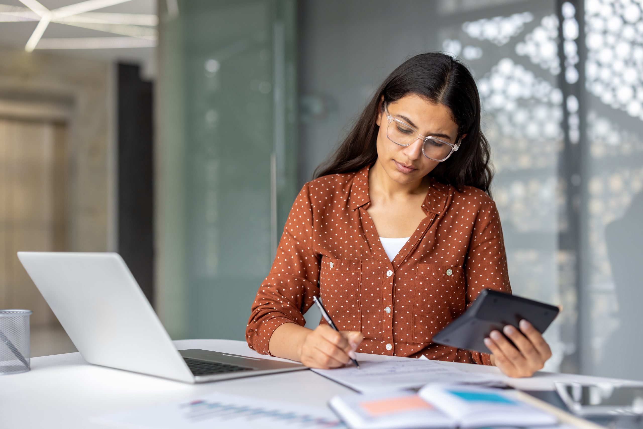 Young,Businesswoman,Calculating,Finances,On,A,Calculator,,Writing,Notes,On